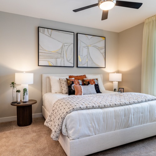 carpeted bedroom with a ceiling fan and large window furnished with a bed, nightstands, and wall art